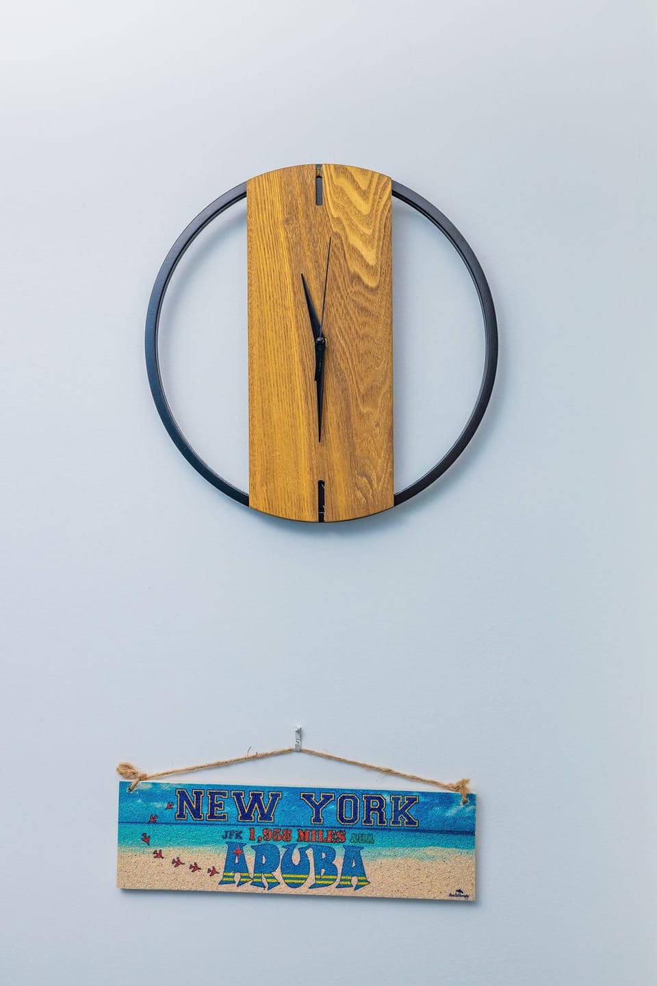 Decorative wall clock with wood grain detail sits above a vintage-style sign marking distance from New York to Aruba—a playful reminder to leave everyday worries behind.