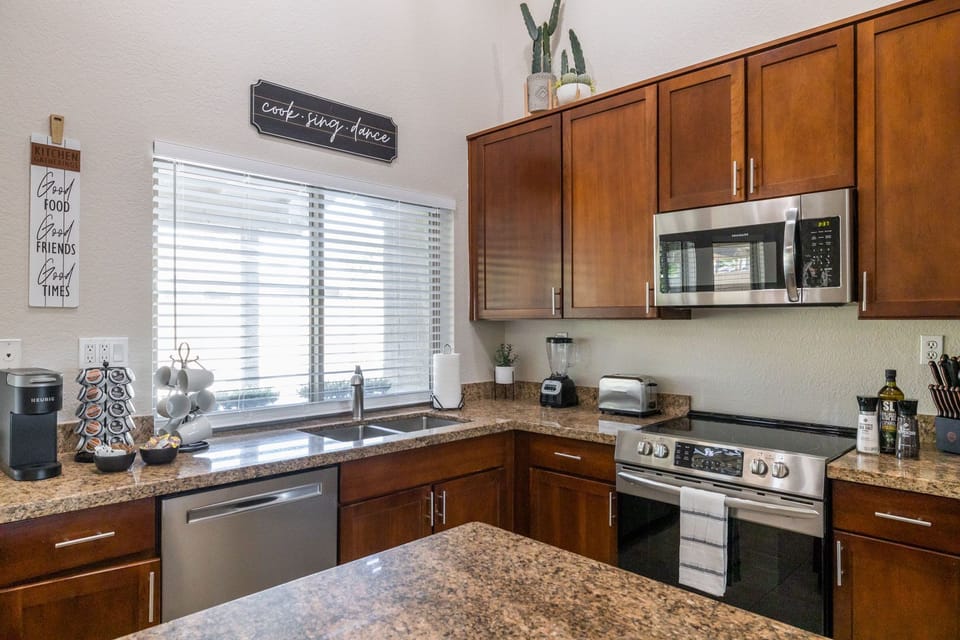 A modern kitchen with wooden cabinets, granite countertops, a stainless steel stove, microwave, and dishwasher. A window with blinds is above the sink. Decor items and small appliances are on the counter.
