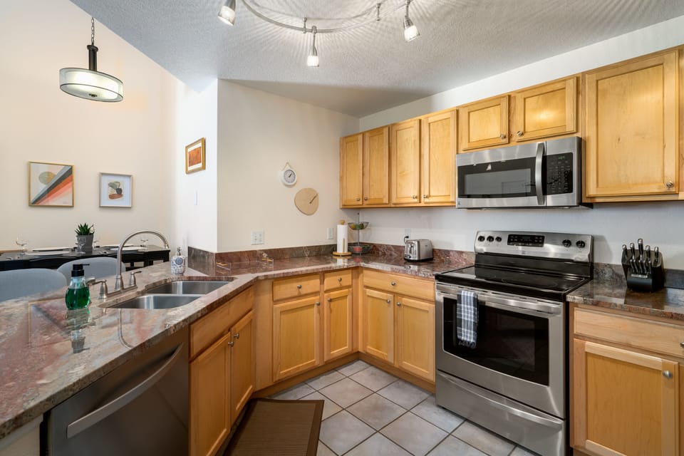 A modern kitchen with wooden cabinets, stainless steel appliances, granite countertops, a double sink, and tiled flooring. Adjacent dining area is visible in the background.