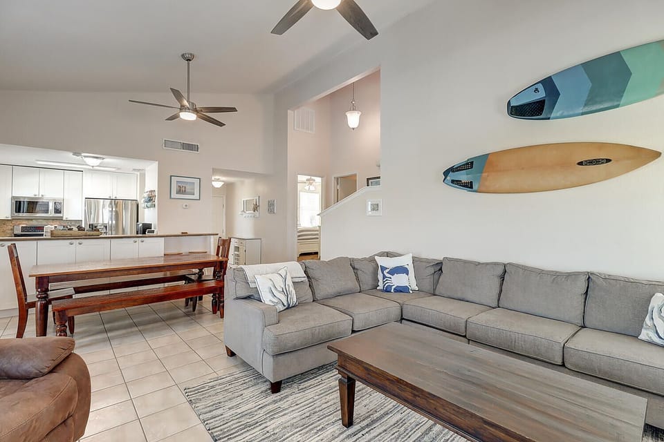 Relax in your open-concept living space with plush sectional seating, coastal surfboard art, and modern kitchen nearby.