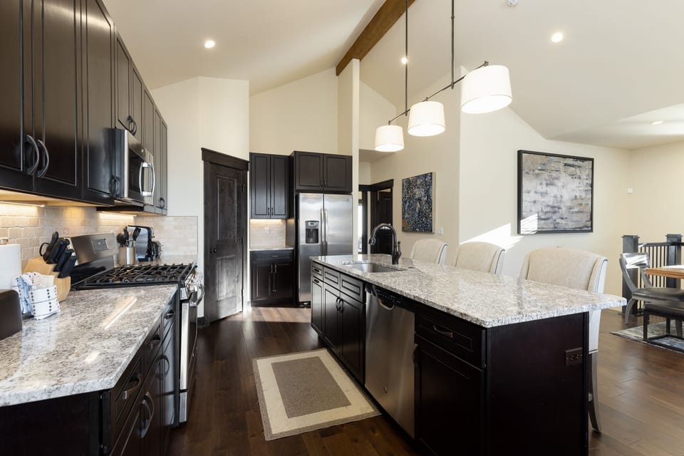 Modern kitchen with dark wood cabinets, stainless steel appliances, granite countertops, and hanging pendant lights. A large island with seating faces the open dining area.