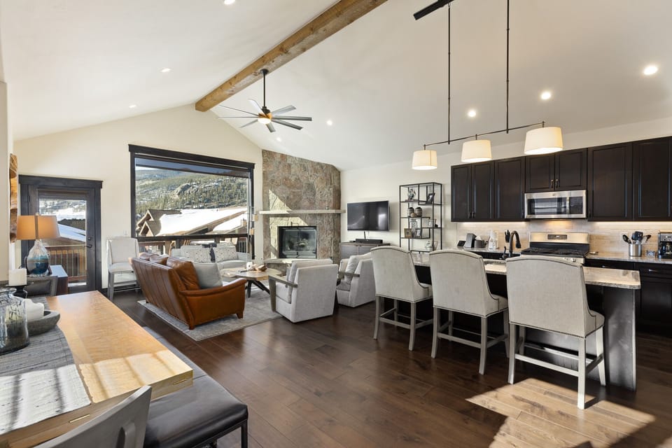 Spacious modern living room and kitchen with vaulted ceiling, wooden beams, large windows, leather sofa, stone fireplace, island with barstools, and dark cabinetry. Scenic view of mountains outside.