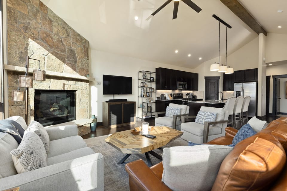 A modern living room with a stone fireplace, flat-screen TV, and a mix of leather and fabric furniture. The open kitchen features dark cabinetry, a breakfast bar with stools, and pendant lighting.