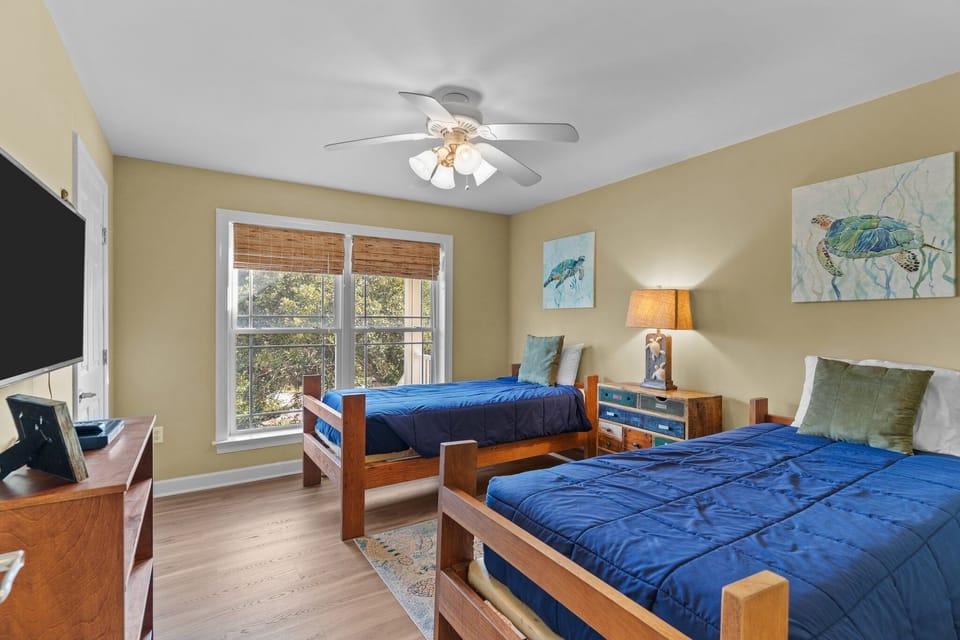 Bright guest bedroom featuring two twin beds with blue bedding, coastal turtle artwork, nightstand with lamp, dresser with TV, and large window bringing in natural light in this Oak Island beach home.