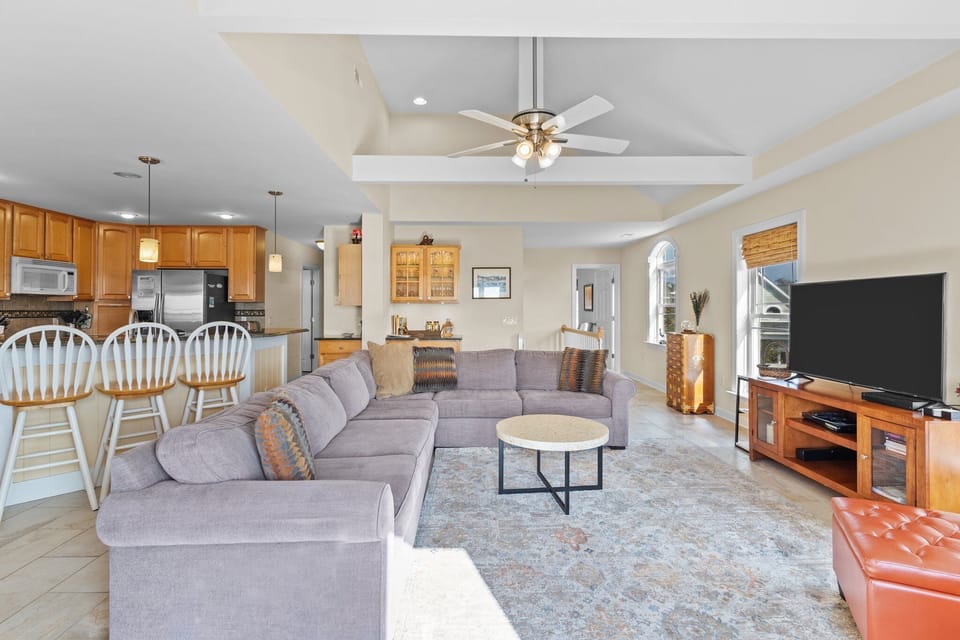 Spacious open-concept living area with vaulted ceilings, sectional sofa, fireplace, armchairs, and large rug flowing into the dining area and kitchen with bar seating. Glass doors lead to a deck with beautiful ocean views in Oak Island, NC
