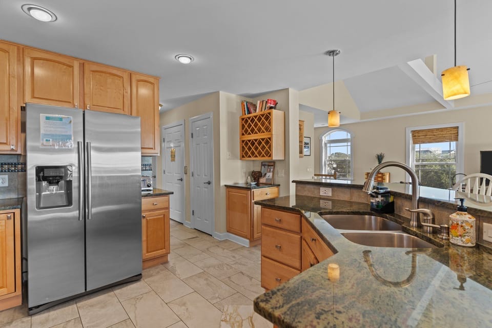 Bright kitchen with natural wood cabinets, granite countertops, stainless refrigerator and dishwasher, electric range, microwave, and small appliances. Window above the sink brings in natural light in this Oak Island beach home.