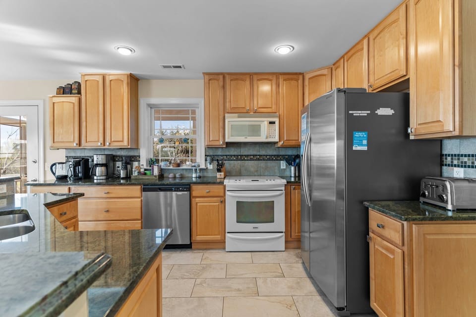 Bright kitchen with natural wood cabinets, granite countertops, stainless refrigerator and dishwasher, electric range, microwave, and small appliances. Window above the sink brings in natural light in this Oak Island beach home.