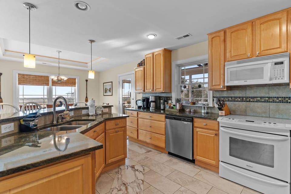 Bright kitchen with natural wood cabinets, granite countertops, stainless refrigerator and dishwasher, electric range, microwave, and small appliances. Window above the sink brings in natural light in this Oak Island beach home.