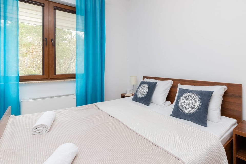 A bedroom with a neatly made bed with blue and white pillows. Blue curtains provide contrast against the natural light entering through the window.