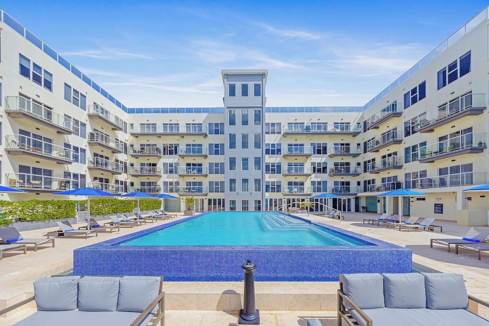 A sun-soaked courtyard pool stretches before modern terraced balconies, offering a tranquil retreat with ample lounge seating and shaded umbrellas beneath clear blue skies.
