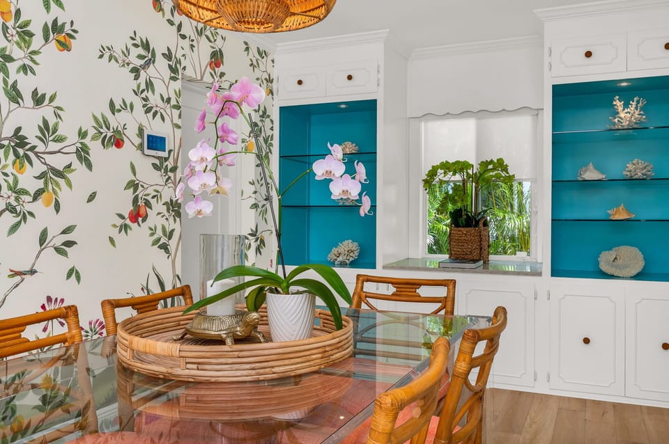 The dining area radiates distinctive charm, adorned with vibrant wallpaper and captivating artwork that imbue the space with charisma. A timeless Palm Beach dining table comfortably accommodates six guests and provides seamless access to the outdoors.