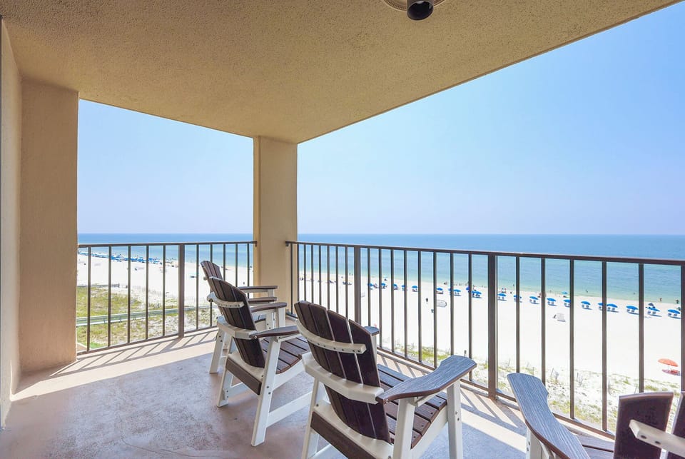 The balcony features two wooden chairs overlooking a serene beach with white sand and blue umbrellas lining the shore
