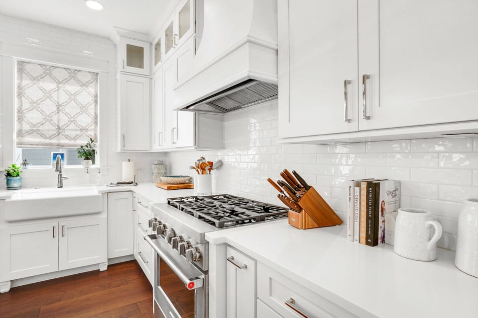 Your pristine white kitchen awaits with gleaming countertops, professional gas range, and thoughtful touches like fresh cookbooks and herbs by the window.
