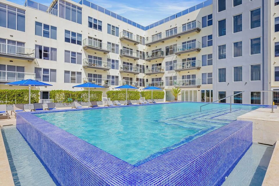 Modern apartment complex featuring a striking glass-walled pool in an inner courtyard. The pool area is surrounded by a well-maintained landscaped hedge, vibrant blue umbrellas, and loungers—ideal for relaxation under sunny skies.