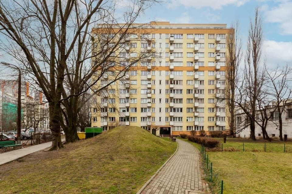 A pathway leading to a residential building surrounded by a grassy area, with trees and benches adding a welcoming outdoor environment.