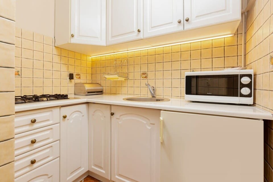 A compact kitchen with white cabinets, a tiled backsplash, a microwave, and bright under-cabinet lighting creating a welcoming atmosphere.