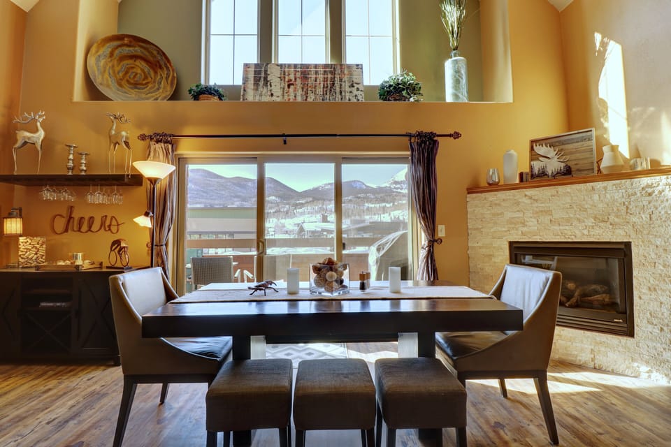 A dining room with a table and four chairs, large windows, a fireplace, and various decor items in a cabin-style interior with mountain views outside the window.