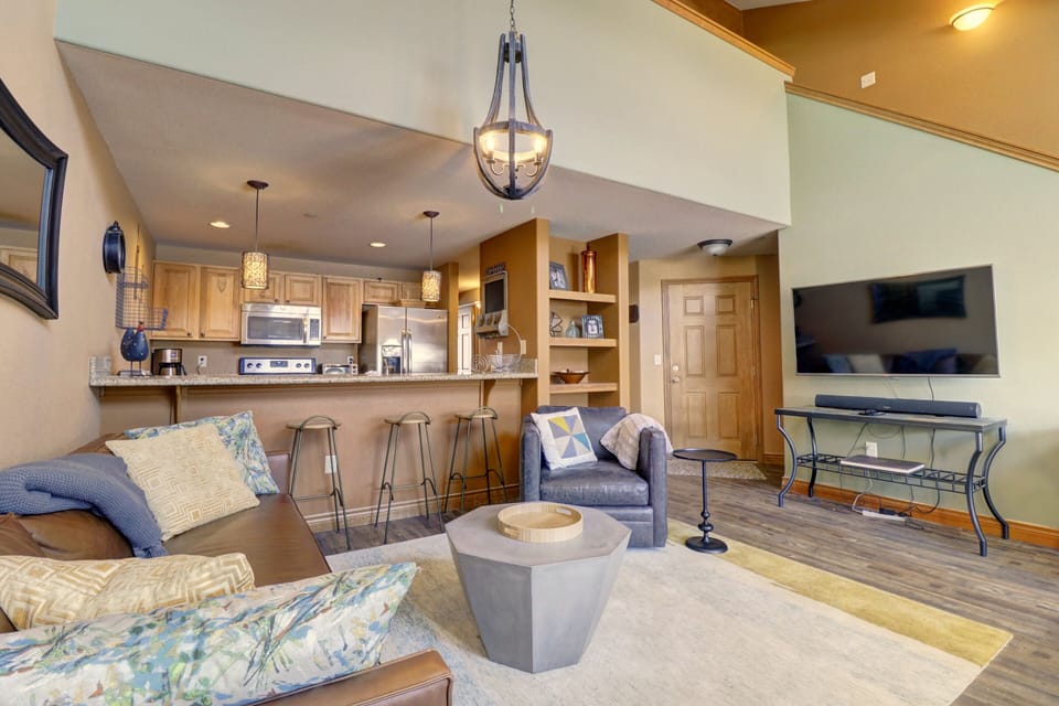 A modern living room with a high ceiling, featuring a brown sofa, a grey armchair, a TV mounted on the wall, a dining bar with bar stools, and a kitchenette with wooden cabinets and stainless steel appliances.