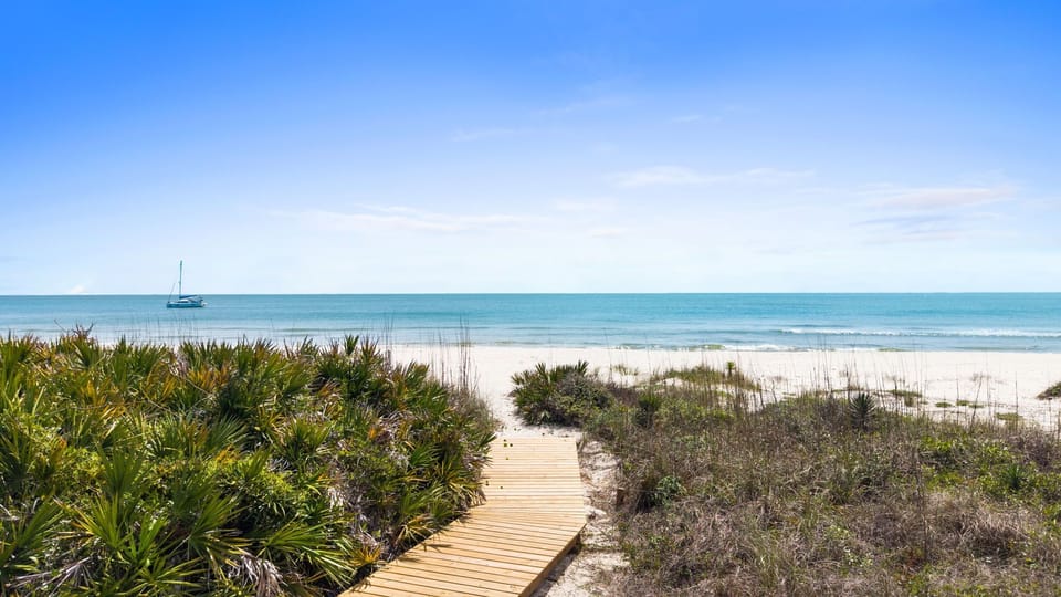Pristine beach with wooden boardwalk leading through coastal vegetation to turquoise waters and white sand.