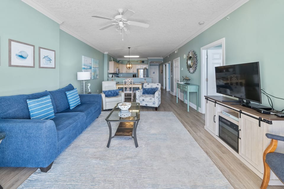 Relax in your coastal-inspired living room with plush blue sofa, entertainment center, and seamless flow to your fully equipped kitchen.