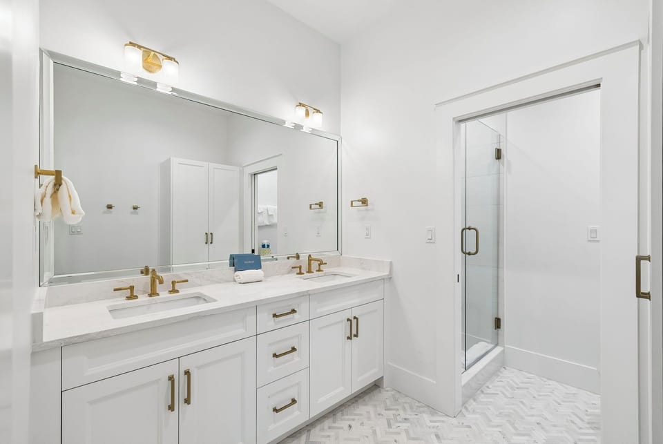 Step into your pristine bathroom where dual vanities with marble countertops and warm brass fixtures frame your morning routine, while the spacious glass-enclosed shower awaits your refreshing escape.