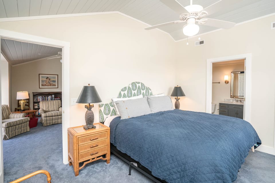 Large bedroom with king bed and navy quilt, vaulted ceiling with fan, window with curtains, wicker dresser with mirror and lamp, soft carpet, and doorway leading to private bathroom in this Oak Island beach house.