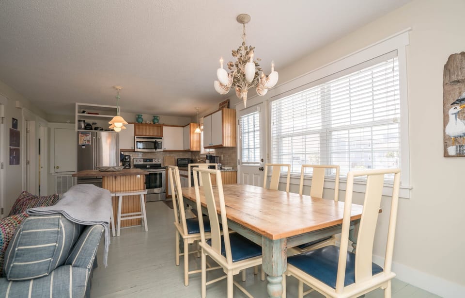 Bright dining area with wooden table, six chairs, shell-style chandelier, large windows with blinds, and open view to kitchen with island seating, stainless appliances, and pendant lighting in this Oak Island beach house.