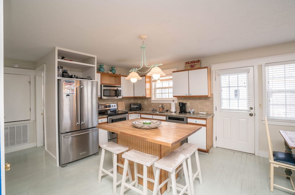 Bright kitchen with stainless refrigerator, stove, microwave, dishwasher, granite countertops, wood island with three stools, pendant lighting, coffee maker, white cabinets, and door leading outside in this Oak Island beach house.