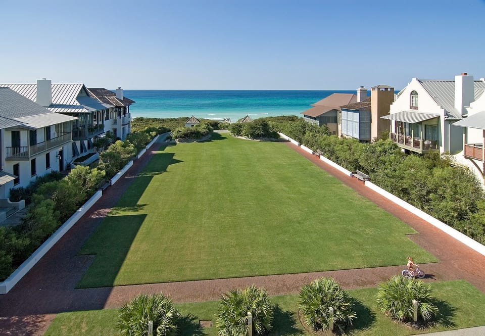 Coastal vacation community with direct beach access, manicured lawns, and charming residential buildings framing a central green space under clear blue skies.