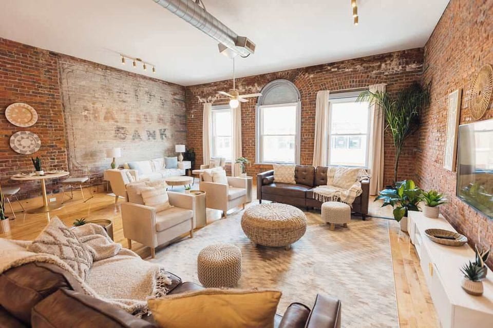 Bright, thoughtfully designed loft in downtown Johnson City featuring exposed brick walls, warm wood floors, and large windows. A cozy seating area with woven chairs and soft textiles sits beneath a vintage mural, blending historic charm with modern 