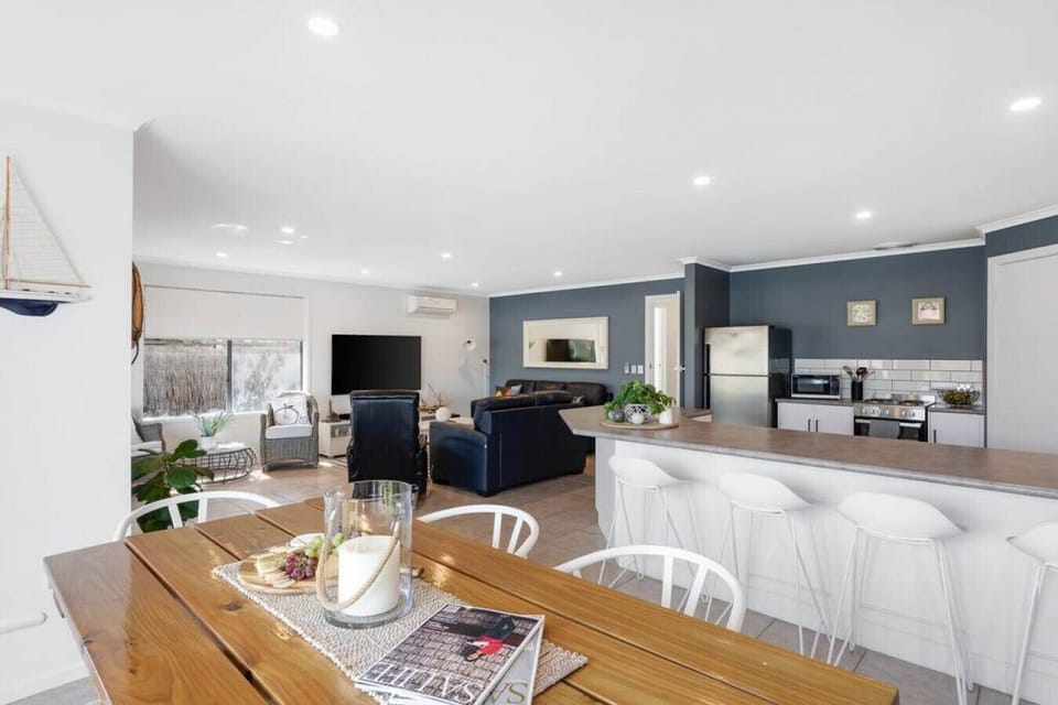 This open-plan living space combines a cozy lounge, modern kitchen, and dining area in a bright, coastal-inspired design. The kitchen features sleek appliances, a breakfast bar with white stools, and blue-gray walls with white tile accents. The adjacent l