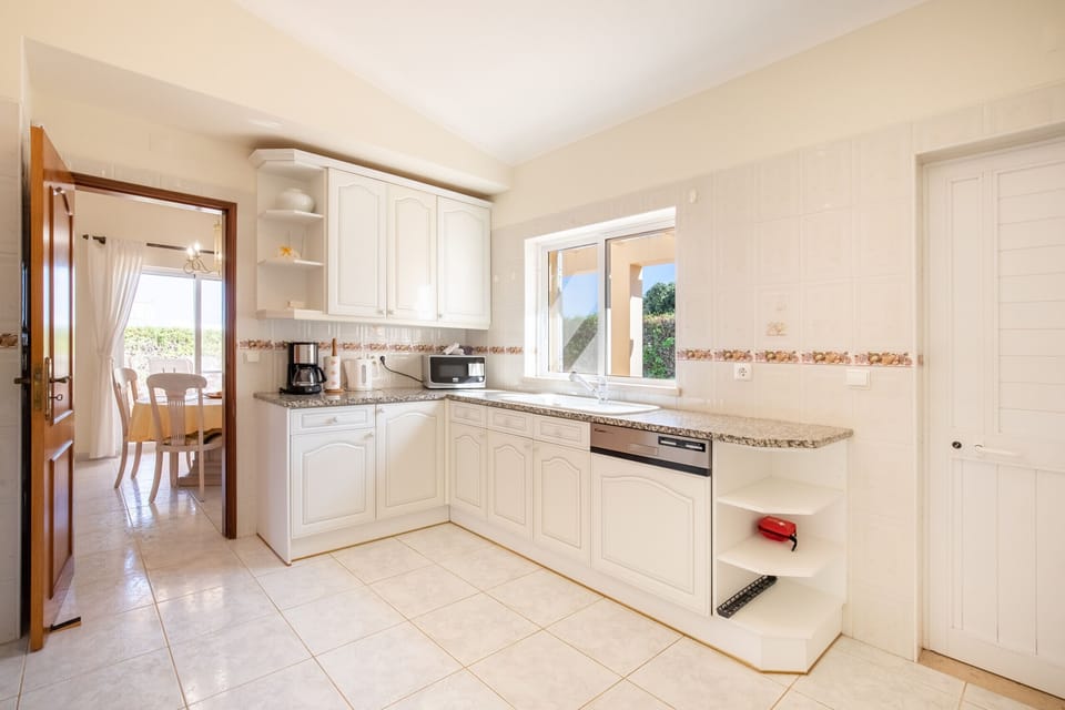 This image shows a modern and bright kitchen with white cabinets and plenty of counter space. A large window above the sink lets in lots of natural light. The kitchen has an open walkway leading to a dining area, which makes it very functional and inviting.