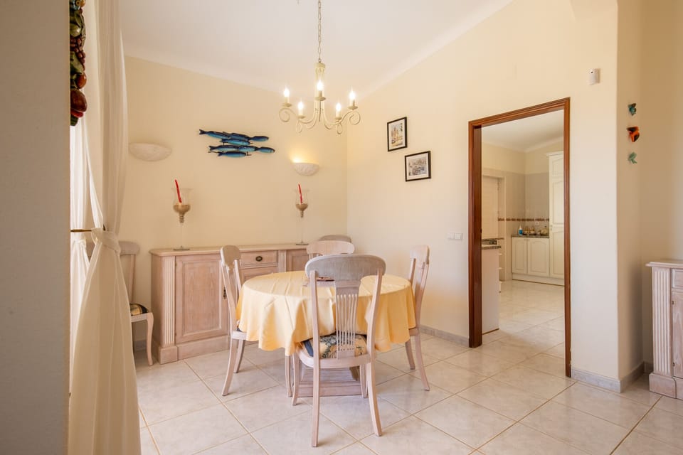 This image shows a bright and inviting interior connecting the dining and living areas. A beautifully set dining table sits near a large patio door, which lets in plenty of sunlight and provides a view of the pool and terrace. The room is comfortably furnished and provides an easy transition to the outdoor area.