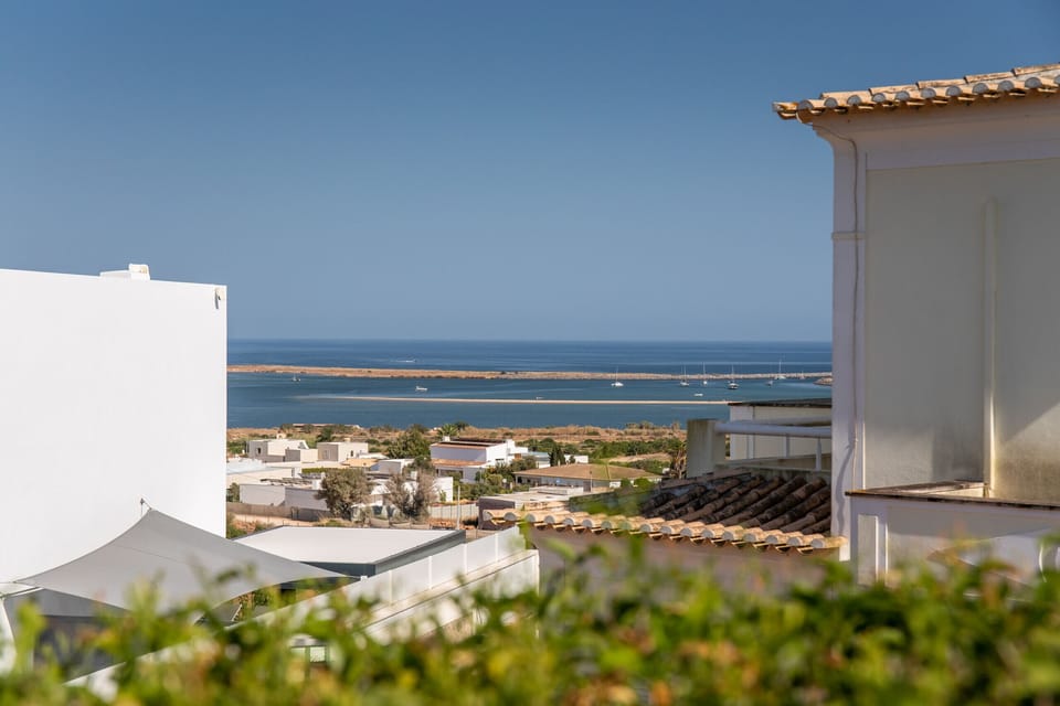 This image shows a great sea view from a balcony. In the foreground, parts of a white building and a terrace with a sunshade hint at a beautiful vista. In the background, a peaceful coastal town and a tranquil bay with a few boats invite relaxation.