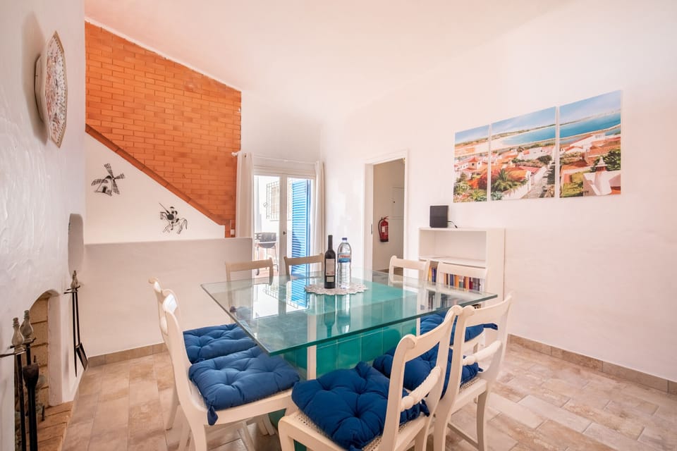 This photo shows another view of the accommodation's dining area. The room is equipped with a glass-top table and white chairs with blue seat cushions. A prominent feature is the exposed brick wall leading to the stairs. Above a low console, three pictures with coastal motifs are hanging.