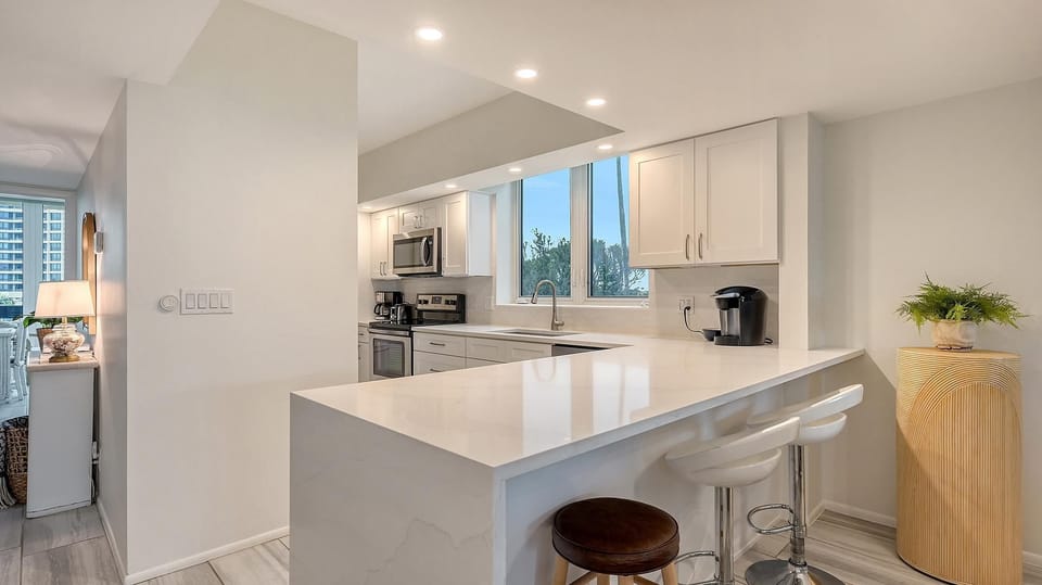 White quartz peninsula with three bar stools extends from the kitchen featuring stainless steel appliances and shaker cabinets.
