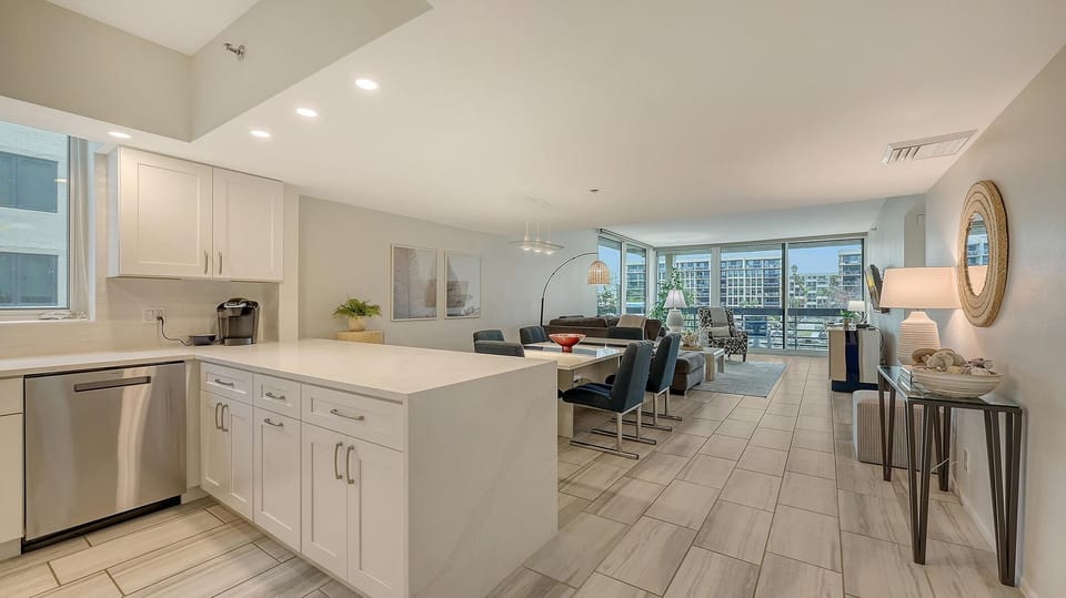 Open-concept kitchen and dining area with white cabinetry, stainless steel dishwasher, and navy upholstered chairs.