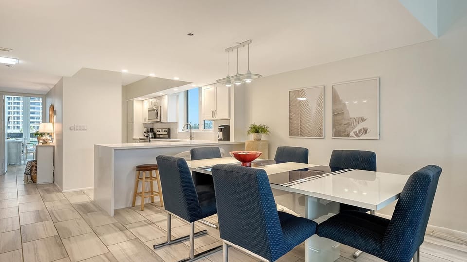 Glossy white dining table extends beneath a modern pendant light, surrounded by six navy upholstered chairs.