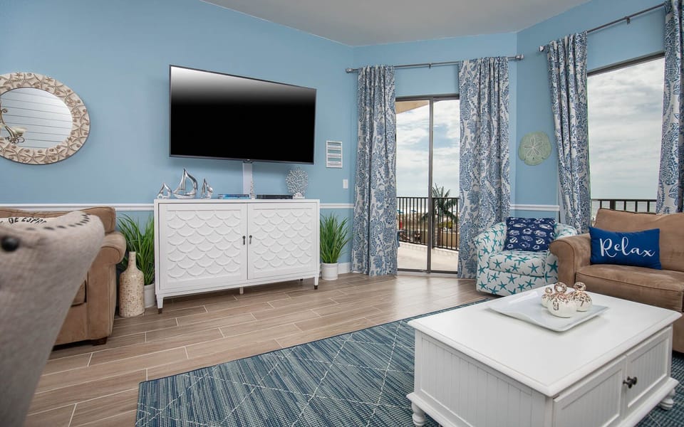 The living area features a cozy sofa with queen sleeper, blue accents, and large windows that invite in the beach views