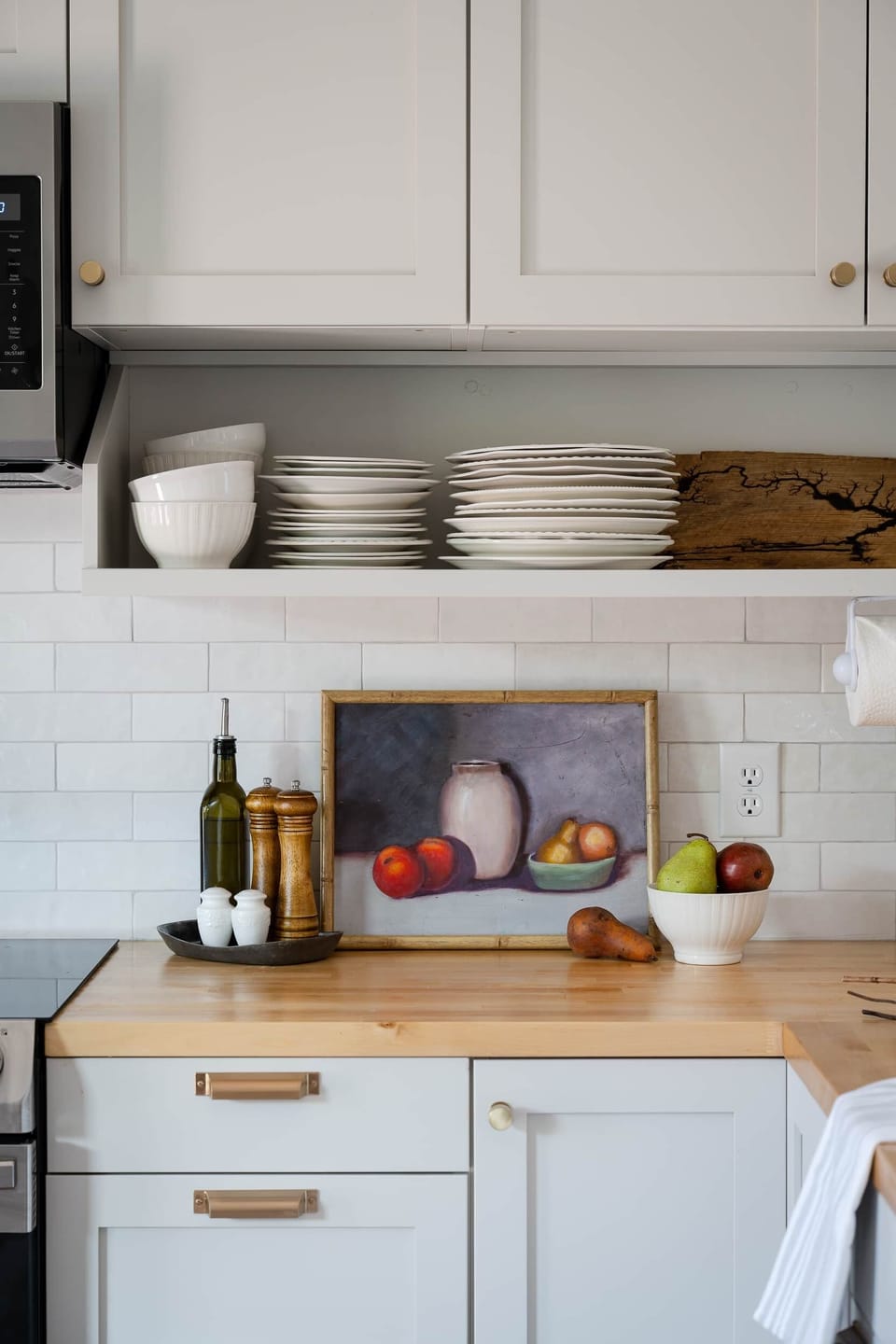 3-East kitchen counter with stacked dishware, fresh fruit, and a still life painting.