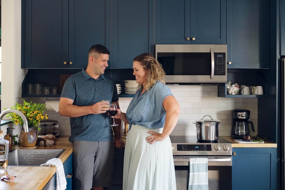 A man and woman share drinks and conversation in a kitchen with blue cabinets and stove.