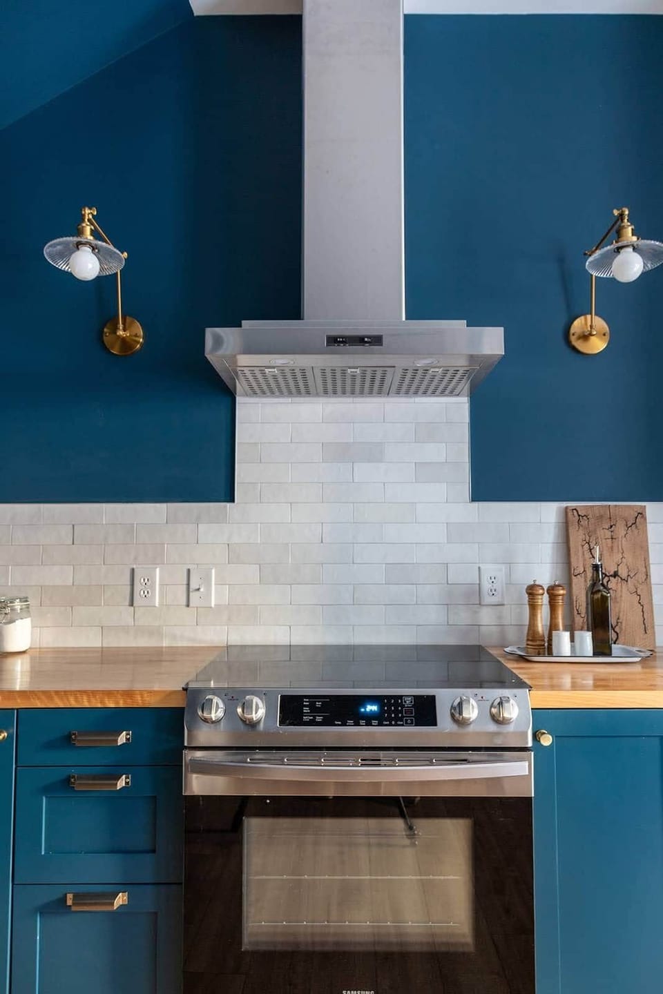 Stainless range and hood centered under brass sconces with white tile and wood countertops.