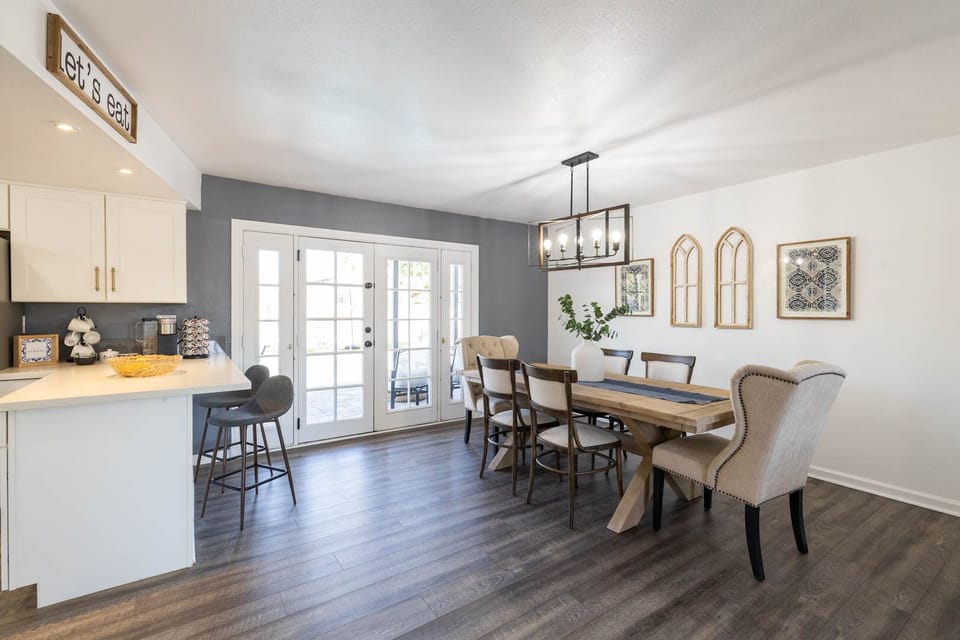 A dining area with a wooden table and six chairs, adjacent to a kitchen counter with stools. French doors open to the outside. The walls have framed art, and the floor is wooden.