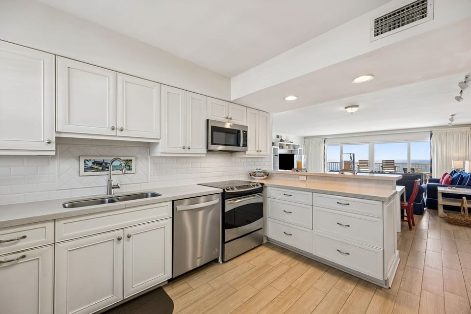 Step into your bright, fully-equipped kitchen with crisp white cabinetry, stainless steel appliances, and ocean views just beyond the open living area.