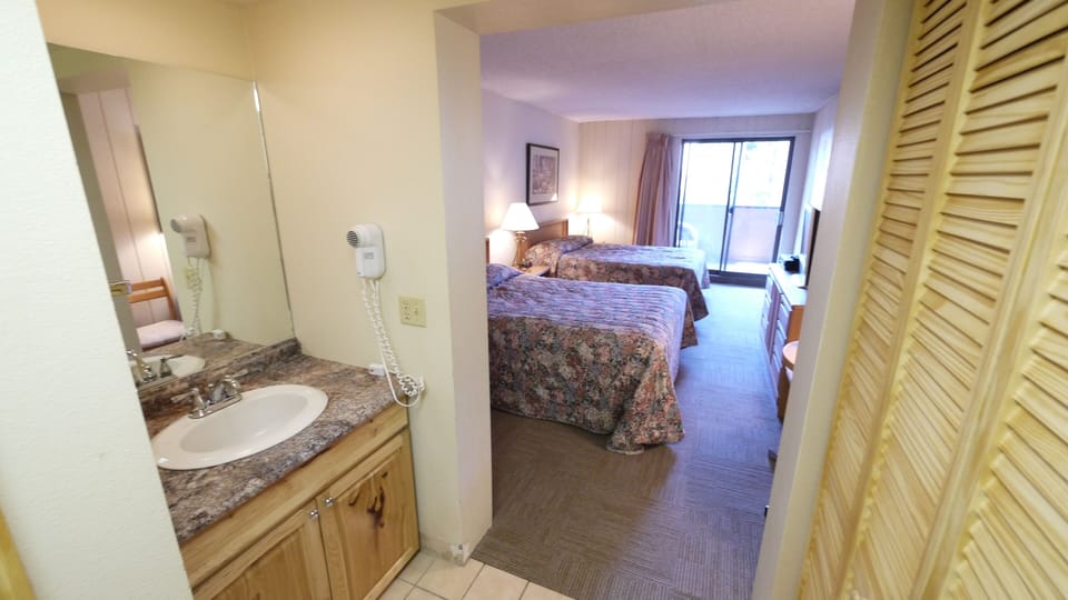 Hotel room with two beds, a television, a small table, and sliding glass doors leading to a balcony. In the foreground, there is a bathroom sink with a wall-mounted hairdryer.
