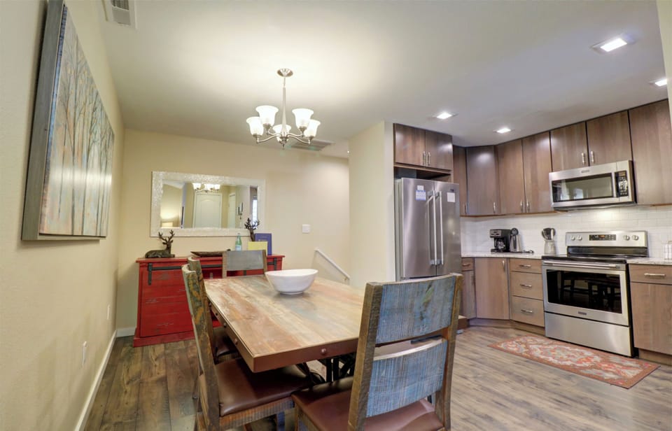 A modern kitchen and dining area featuring a wooden dining table with colorful chairs, stainless steel appliances, wooden cabinets, a red side table, and a wall mirror.