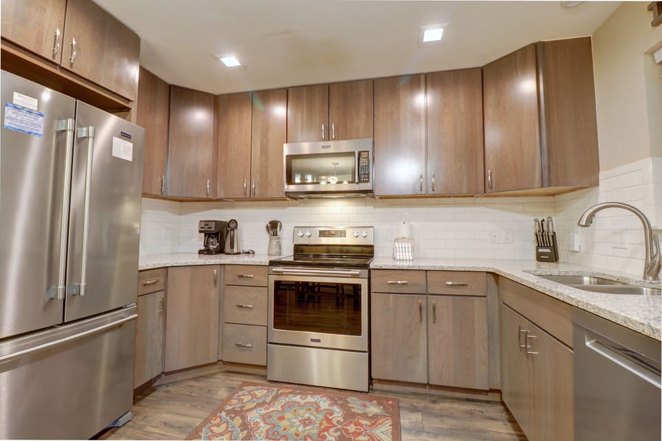 A modern kitchen with stainless steel appliances, including a refrigerator, microwave, and stove, brown wooden cabinets, a coffee maker, white subway tile backsplash, granite countertops, and a patterned rug.