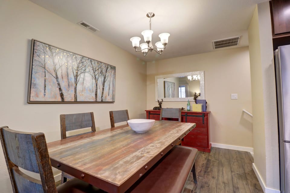 A dining room with a wooden table, six chairs, a red sideboard with decorative items, a framed tree artwork, and a rectangular mirror on the wall. A chandelier hangs above the table.
