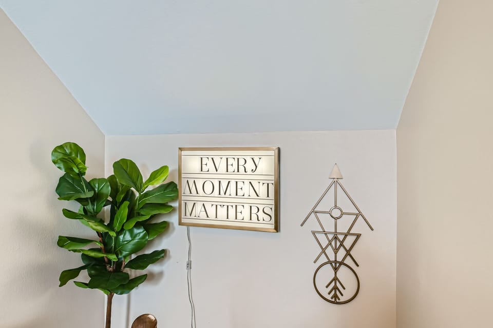 A sign reading "Every Moment Matters" is mounted on a white wall above a decorative metal wall art piece and a large green potted plant.