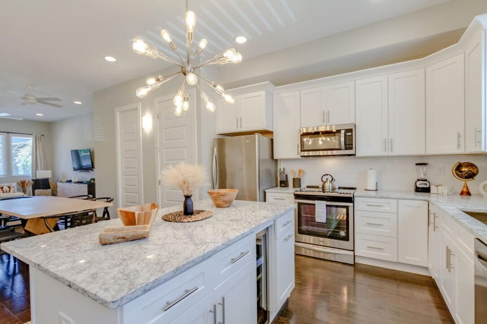 Full kitchen view showcasing white cabinets, stainless steel appliances, a marble island, and warm hardwood floors.